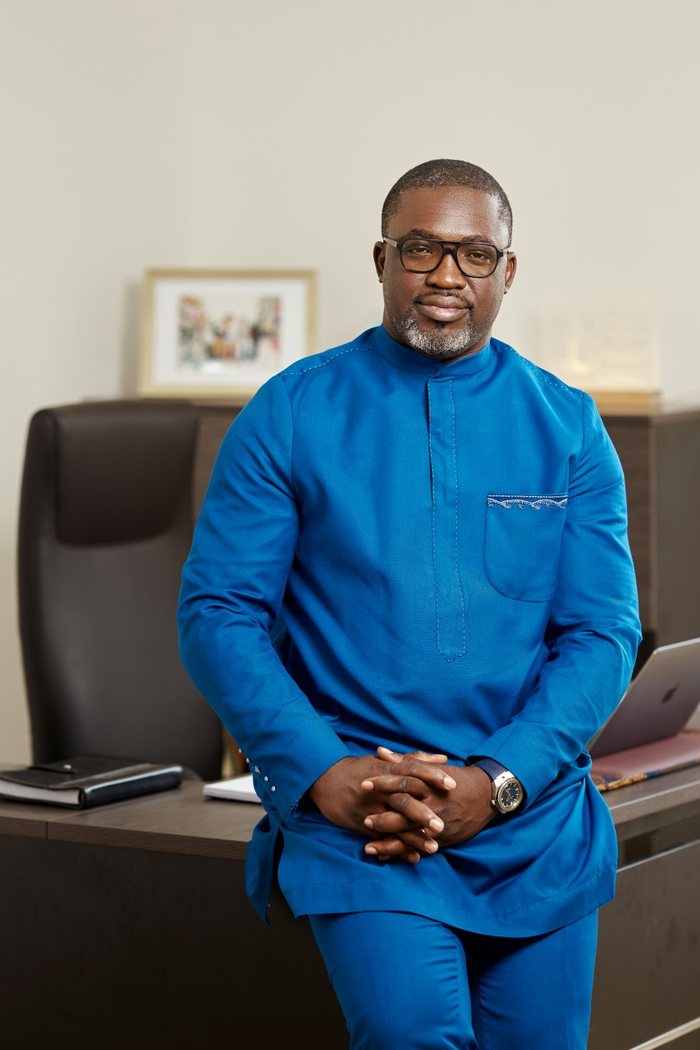 Rodrigue Mboumba Bissawou in a blue outfit, leaning against his desk, hands clasped