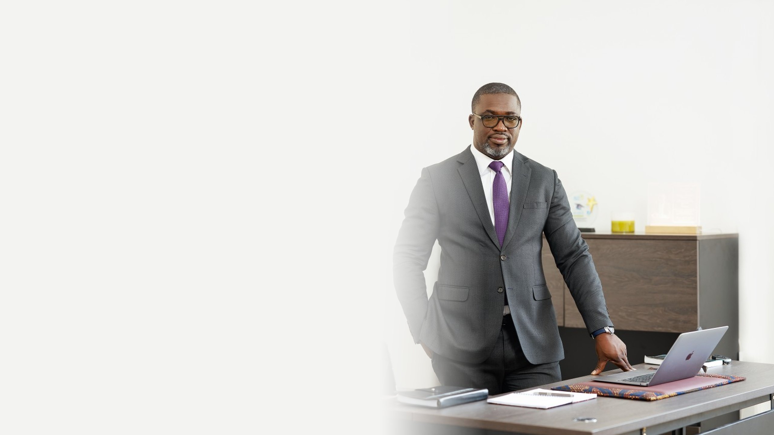 Rodrigue Mboumba Bissawou standing behind his desk in a grey suit and purple tie
